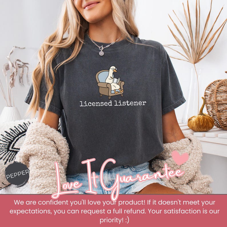 May include: Dark gray t-shirt with the text "licensed listener." The shirt features a cartoon goose wearing glasses and sitting in a chair, reading a book. The shirt is a comfortable, casual style.