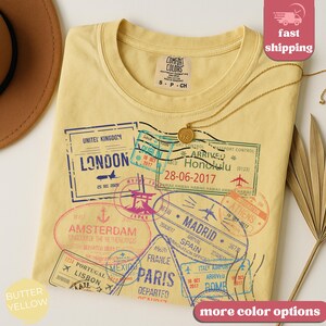 May include: Butter yellow Comfort Colors t-shirt with travel-themed stamp graphics. The stamps include locations like London, Amsterdam, Paris, and Honolulu. The shirt has a crew neck and a small tag that says "S-P-CH".