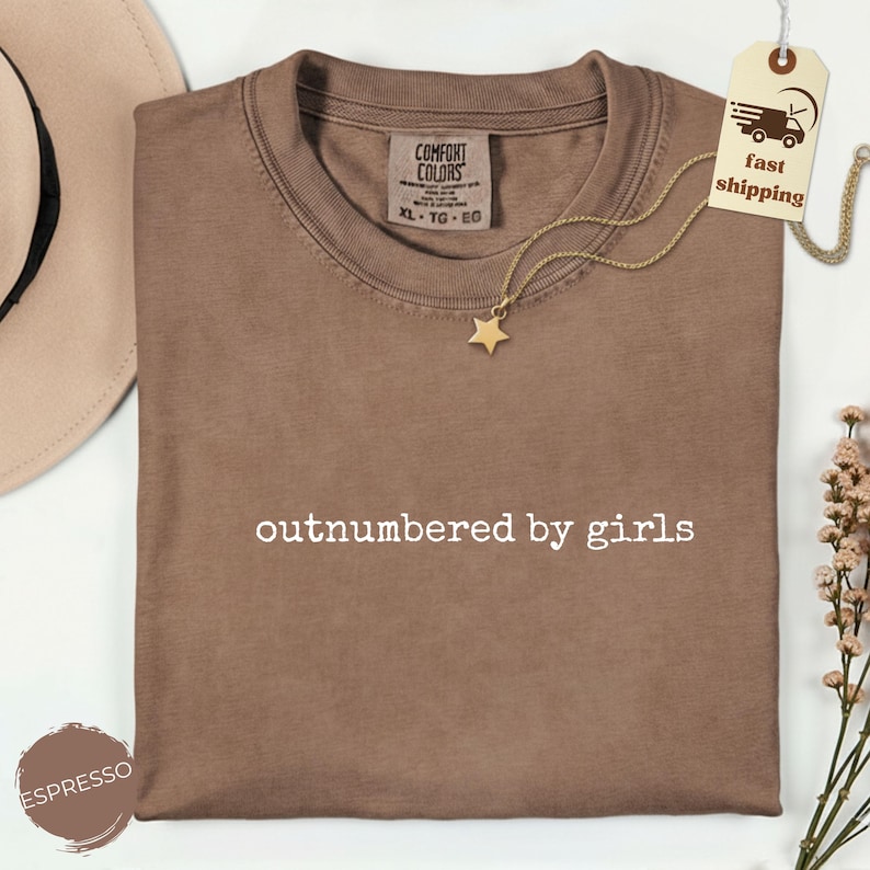 May include: Espresso-colored t-shirt with the text "outnumbered by girls" in white. A gold star pendant necklace rests on the shirt. The shirt has a Comfort Colors tag. A fast shipping tag is attached.