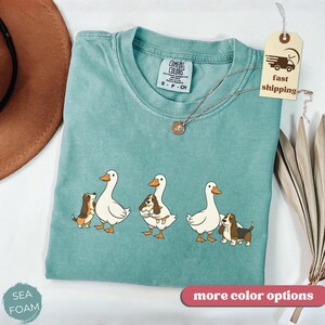 May include: Seafoam green Comfort Colors t-shirt featuring a graphic of three white ducks with orange beaks and feet, each paired with a brown and white dog. The shirt has a crew neck and a small tag that says "S P CH". A gold necklace is draped across the shirt.