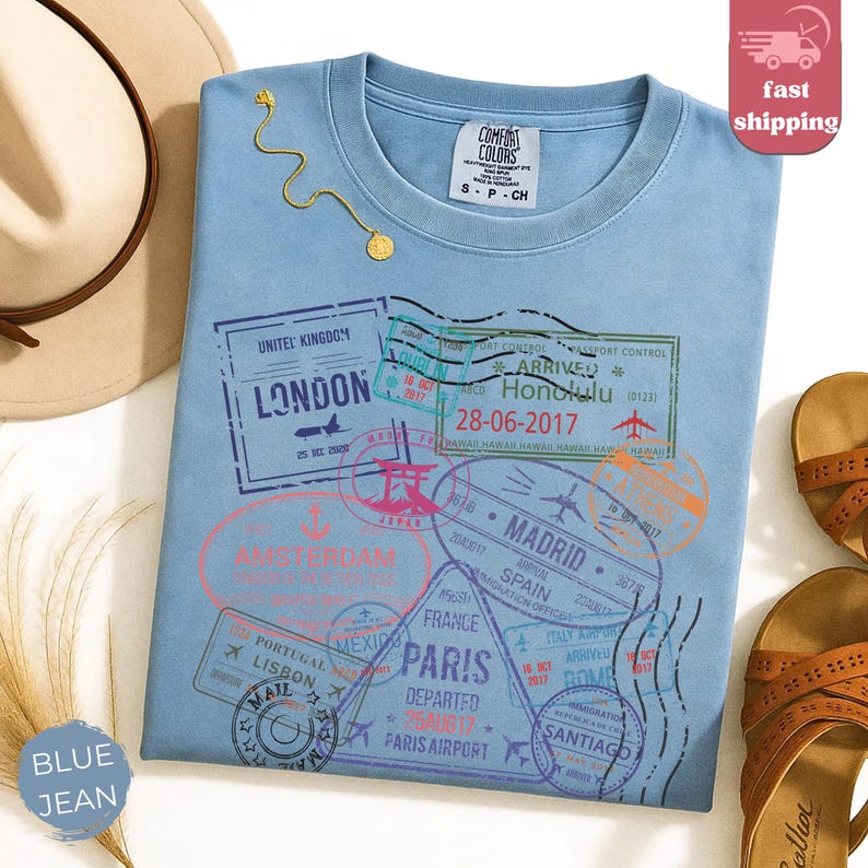 May include: A light blue Comfort Colors t-shirt with travel-themed stamps, including London, Amsterdam, and Paris. The shirt has a small tag at the neckline and a gold necklace. The shirt is labeled "Blue Jean".