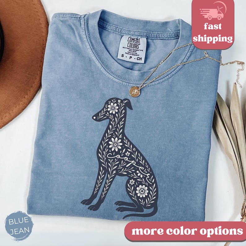 May include: A blue jean t-shirt with a floral design of a greyhound dog. The dog is outlined in black and the flowers are white. The text "Cooper" is spelled out in the flowers on the dog's body.