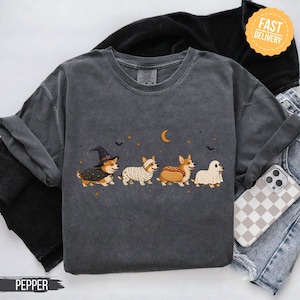 May include: A charcoal grey crew neck t-shirt featuring a graphic of four Corgi dogs in Halloween costumes. The dogs are dressed as a witch, a mummy, a hot dog, and a ghost, set against a background of small orange stars, bats, and a crescent moon.