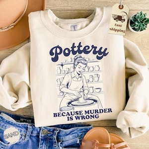 May include: Cream-colored sweatshirt with the word "Pottery" in navy blue script above a black and white illustration of a potter at work. The text "BECAUSE MURDER IS WRONG" is below the illustration.