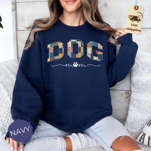 May include: Navy blue sweatshirt with the word "DOG" in a patchwork design. Below the word "DOG" is the word "mom" with a paw print. The sweatshirt is a crew neck style.