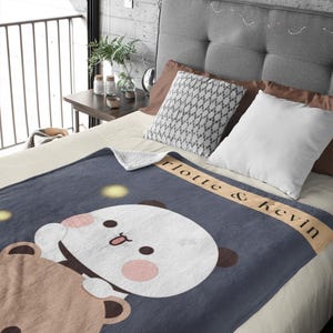 Pu&ograve; includere: Una morbida coperta in pile blu con un orso marrone e un panda bianco che si abbracciano. La coperta ha il testo "Lotte & Kevin" su uno stendardo beige.
