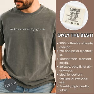 May include: Pepper-colored t-shirt with the phrase "outnumbered by girls" in white. The shirt is made of 100% cotton and has a relaxed fit. The tag reads "Comfort Colors" and "Made in Honduras".