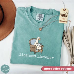 May include: Seafoam green t-shirt with a graphic of a goose wearing glasses and reading in a chair. The text "licensed listener" is printed below the graphic in white. A gold necklace is draped on the shirt. A tag with a fast shipping icon is attached.