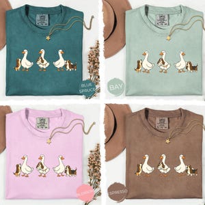 May include: Four Comfort Colors t-shirts in blue spruce, bay, blossom, and espresso. Each shirt features a graphic of three white geese and three brown and white dogs. Each shirt has a gold star necklace.