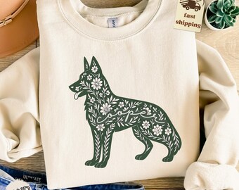 May include: Cream-colored sweatshirt featuring a dark green floral dog design with the word "Cooper" in cursive. The dog silhouette is filled with flowers and leaves. The sweatshirt has a crew neck and long sleeves.