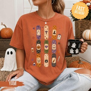 May include: A burnt orange short-sleeve t-shirt featuring a graphic print of nine vertical adhesive bandages decorated with various Halloween and medical icons, including a ghost, pumpkin, skull, and stethoscope, alongside small scattered motifs.