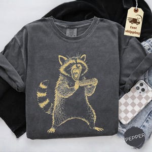 May include: Dark grey long-sleeve shirt featuring a yellow illustration of a raccoon playing a harmonica. The raccoon is depicted with an open mouth and a fierce expression. The shirt is made of a soft, comfortable material.