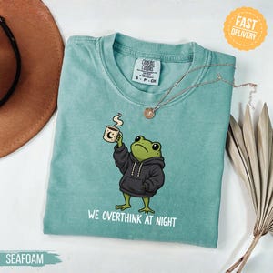 May include: A seafoam green t-shirt featuring a graphic of a green frog wearing a black hoodie and holding a steaming mug. The text below the frog reads, "WE OVERTHINK AT NIGHT." The shirt has a Comfort Colors brand label at the inner neckline.