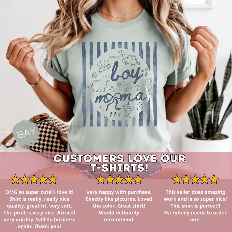 May include: A light green t-shirt with a blue and white design. The design includes the words "boy momma" in a circle with illustrations of a dinosaur, a robot, and other toys. The shirt is a crew neck.