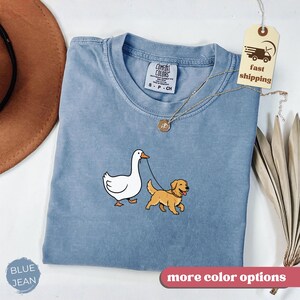 Pu&ograve; includere: T-shirt Comfort Colors blu jeans con grafica di un'oca bianca che porta a guinzaglio un golden retriever. La maglietta ha girocollo e maniche corte. L'etichetta recita "fast shipping".