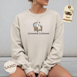 May include: Sand-colored sweatshirt with the text "licensed listener" below a cartoon goose in a chair reading a book. The goose is wearing glasses. A fast shipping tag is attached to the sweatshirt.