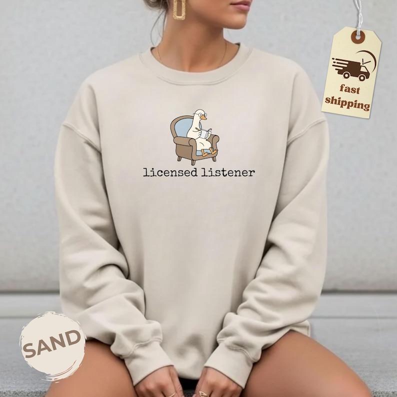 May include: Sand-colored sweatshirt with the text "licensed listener" below a cartoon goose in a chair reading a book. The goose is wearing glasses. A fast shipping tag is attached to the sweatshirt.