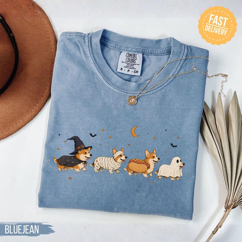 May include: A blue crew neck t-shirt featuring a graphic of four Corgi dogs in Halloween costumes, including a witch, a mummy, a hot dog, and a ghost. The shirt has a Comfort Colors label at the neck and is decorated with stars, a moon, and bats.