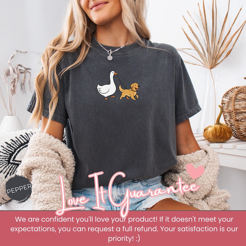 Pu&ograve; includere: T-shirt grigio scuro con un cartone animato di un'oca che porta a spasso un golden retriever al guinzaglio. La maglietta ha un girocollo e maniche corte. Il testo "Love It Guarantee" &egrave; stampato sulla maglietta.