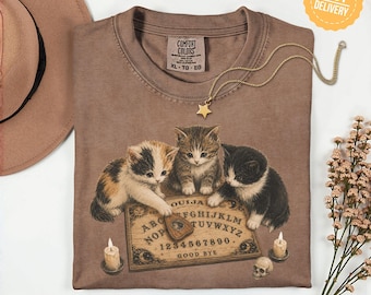 Könnte beinhalten: Ein braunes T-Shirt mit einer Grafik von drei Kätzchen, die um ein Ouija-Brett versammelt sind. Die Kätzchen berühren eine Planchette auf dem Brett, das das Alphabet, Zahlen und die Worte GOOD BYE zeigt. Zwei brennende Kerzen und ein kleiner Totenkopf sind abgebildet.