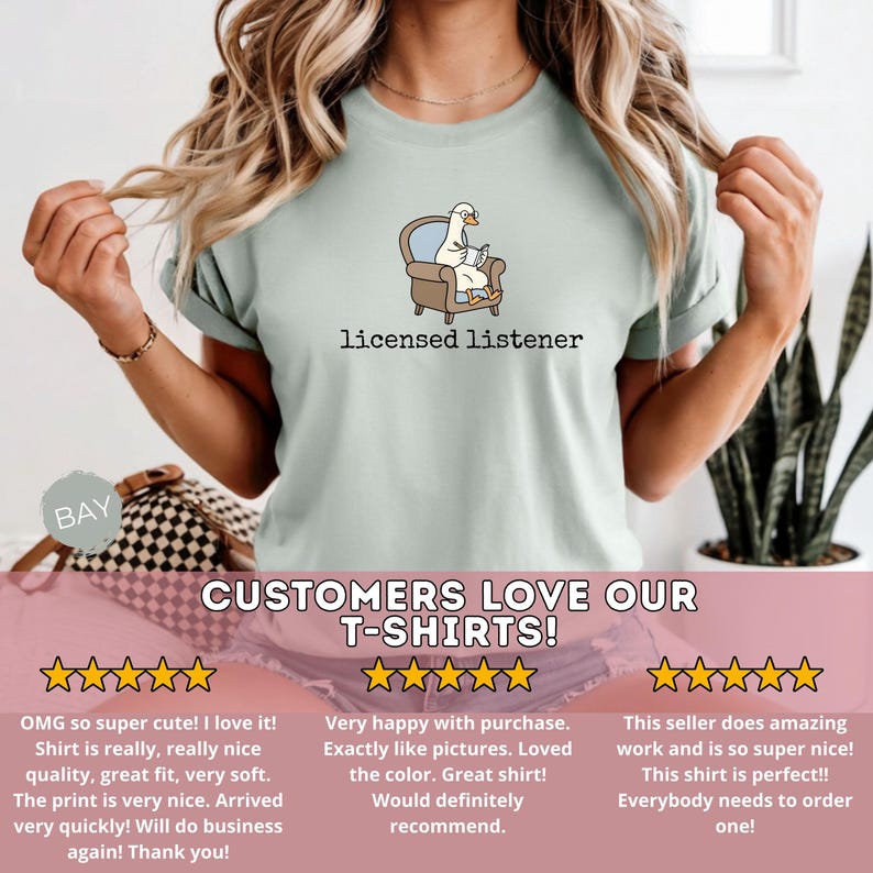May include: A light green t-shirt featuring a cartoon goose in an armchair reading a book, with the text "licensed listener" below. The shirt has a crew neck and short sleeves. The shirt is part of a t-shirt collection.