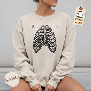 May include: Sand-colored sweatshirt featuring a black and white rib cage graphic. The letters "R" and "L" are printed above the rib cage. A tag with the text "Gildan 18000" is attached to the sweatshirt.