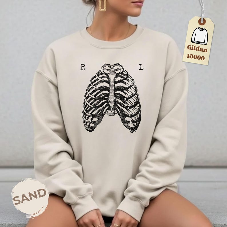 May include: Sand-colored sweatshirt featuring a black and white rib cage graphic. The letters "R" and "L" are printed above the rib cage. A tag with the text "Gildan 18000" is attached to the sweatshirt.