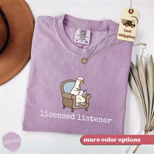 May include: A lilac-colored t-shirt with the text "licensed listener" printed in white. The shirt features a cartoon goose wearing glasses and reading a book while seated in a brown armchair. A silver necklace with a pendant is on the shirt.