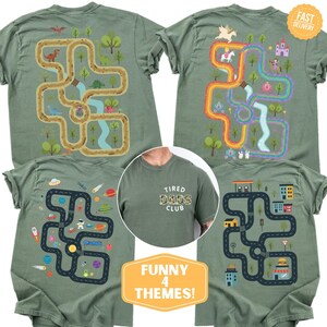 May include: Four olive green t-shirts with different graphic designs. One features a dinosaur maze, another a rainbow unicorn maze, a third a space-themed maze, and the last a city-themed maze. The center shirt has "Tired Dads Club" text.