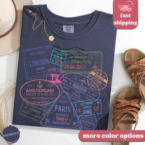 May include: Midnight blue t-shirt with travel-themed stamp graphics, including London, Amsterdam, Paris, and Rome. The shirt features a crew neck and short sleeves. A gold necklace and sandals are also visible.