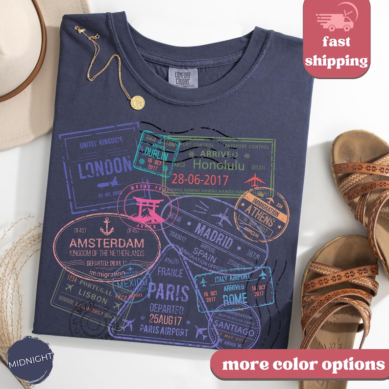 May include: Midnight blue t-shirt with travel-themed stamp graphics, including London, Amsterdam, Paris, and Rome. The shirt features a crew neck and short sleeves. A gold necklace and sandals are also visible.
