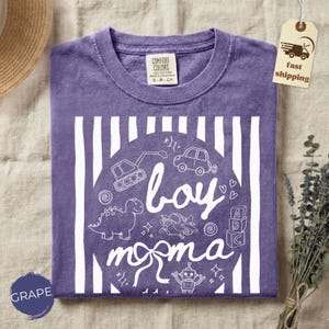 May include: A purple t-shirt with a white graphic that says "boy momma" with illustrations of a dinosaur, car, and other toys. The shirt has a crew neck and short sleeves. The tag says "Comfort Colors".