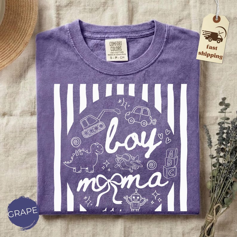 May include: A purple t-shirt with a white graphic that says "boy momma" with illustrations of a dinosaur, car, and other toys. The shirt has a crew neck and short sleeves. The tag says "Comfort Colors".
