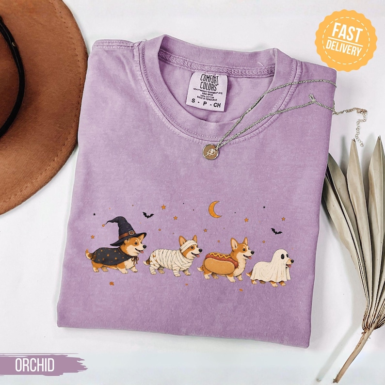 May include: An orchid-coloured t-shirt featuring a graphic of four corgis in Halloween costumes: a witch, a mummy, a hot dog, and a ghost. The design includes a crescent moon, stars, and bats. The shirt has a Comfort Colors label at the neck.