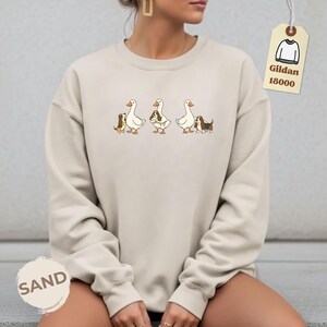 May include: Sand-colored sweatshirt featuring a whimsical design of three geese, each accompanied by a small dog. The dogs and geese are in a cartoon style. The sweatshirt has the word "SAND" printed on it.