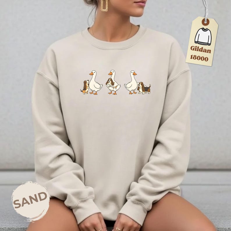 May include: Sand-colored sweatshirt featuring a whimsical design of three geese, each accompanied by a small dog. The dogs and geese are in a cartoon style. The sweatshirt has the word "SAND" printed on it.