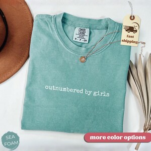 May include: Seafoam green t-shirt with the text "outnumbered by girls" in white. The shirt has a crew neck and short sleeves. A gold necklace with a pendant rests on the shirt. A tag with a fast shipping logo is attached.
