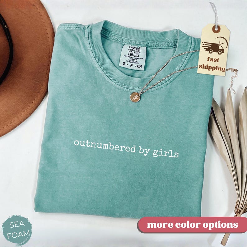 May include: Seafoam green t-shirt with the text "outnumbered by girls" in white. The shirt has a crew neck and short sleeves. A gold necklace with a pendant rests on the shirt. A tag with a fast shipping logo is attached.