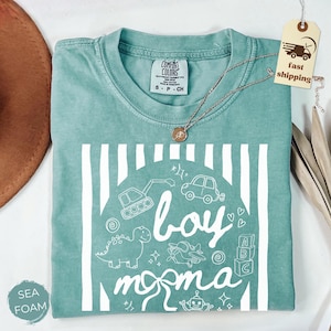 May include: Sea foam green t-shirt with a white graphic design. The design includes the words "boy momma" with illustrations of a dinosaur, car, airplane, and other toys. A silver necklace with a pendant is on the shirt.