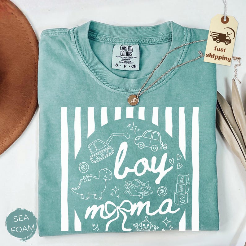 May include: Sea foam green t-shirt with a white graphic design. The design includes the words "boy momma" with illustrations of a dinosaur, car, airplane, and other toys. A silver necklace with a pendant is on the shirt.