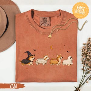 May include: A rust-coloured t-shirt featuring a graphic of four Corgi dogs in Halloween costumes: a witch, a mummy, a hot dog, and a ghost. The design includes small stars, bats, and a crescent moon. The shirt has a Comfort Colors brand label.