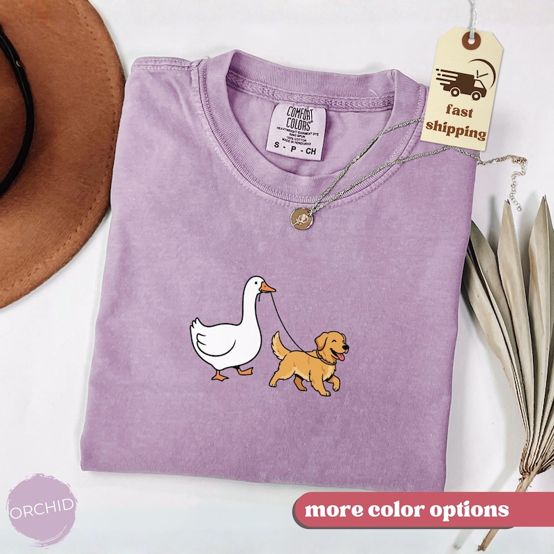 Pu&ograve; includere: Una maglietta lilla Comfort Colors con un design ricamato di un'oca bianca che porta a spasso un golden retriever al guinzaglio. La maglietta ha un girocollo e maniche corte. Una collana d'argento con un ciondolo &egrave; drappeggiata sulla maglietta. L'etichetta dice "spedizione veloce".