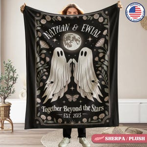 Puede incluir: Manta negra con un diseño que presenta dos figuras de fantasmas blancos, una luna llena y detalles florales. El texto dice "Nathan & Eiwial", "Together Beyond the Stars" y "Est. 2023". La manta está sostenida por una persona.