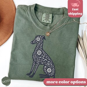 May include: A green t-shirt with a floral design of a greyhound dog. The shirt has a Comfort Colors tag and the text "S - P - CH" on the tag.