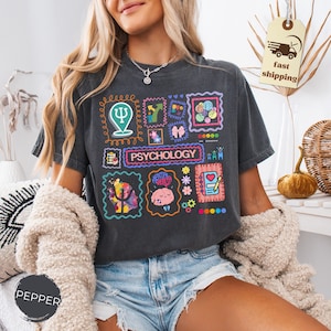 May include: Dark gray t-shirt with a colorful graphic design featuring the word "PSYCHOLOGY" and various psychology-related symbols and illustrations. The design includes a brain, a heart, and other mental health icons.