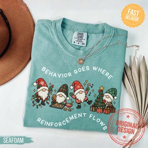 May include: A seafoam green t-shirt featuring a graphic of four gnomes with Christmas decorations and the text "BEHAVIOR GOES WHERE REINFORCEMENT FLOWS". The shirt has a crew neckline and a Comfort Colors brand label inside the collar.