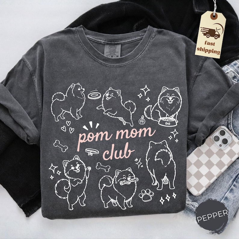 以下が含まれることがあります： 白いポメラニアン犬のグラフィックとピンク色の「pom mom club」の文字が入った、濃い灰色の長袖シャツ。デザインには犬の骨、ハート、足跡が含まれています。シャツはクルーネックです。