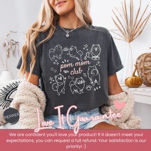 以下が含まれることがあります： ポメラニアン犬の白いグラフィックと「pom mom club」の文字が入った濃い灰色のTシャツ。デザインには犬の骨、星、足跡が含まれています。このTシャツはカジュアルなスタイルです。