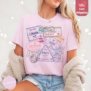 May include: A light pink t-shirt featuring a collection of travel-themed stamps. The stamps include destinations like London, Amsterdam, Paris, and Madrid. The shirt also has the text "Honolulu 28-06-2017".