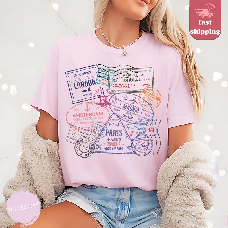 May include: A light pink t-shirt featuring a collection of travel-themed stamps. The stamps include destinations like London, Amsterdam, Paris, and Madrid. The shirt also has the text "Honolulu 28-06-2017".